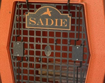 May include: An orange Ruff Land Performance Kennels dog crate with a black grid door. A black rectangular nameplate with an orange pheasant silhouette and the name SADIE in orange text is attached to the center of the door. The crate has a textured finish.