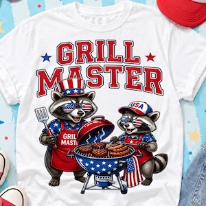 Pode incluir: Camiseta branca com um gráfico de dois guaxinins grelhando. O texto "GRILL MASTER" é em vermelho e branco. Os guaxinins usam óculos de sol, aventais e chapéus com desenhos da bandeira americana. Uma churrasqueira azul e vermelha está no centro.