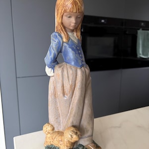 Può includere: Una statuetta in ceramica raffigurante una persona con lunghi capelli castano-rossicci e un fiocco bianco, che indossa una giacca blu e una lunga gonna color marrone chiaro. La figura poggia su una base verde accanto a un cagnolino.