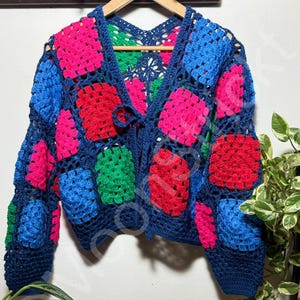 May include: A colorful, handmade crochet cardigan. The open-front sweater features a navy blue base with vibrant squares of pink, red, green, and blue. The cardigan is displayed on a wooden hanger against a white wall.