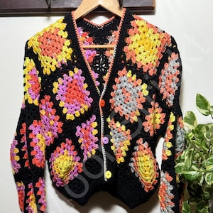 Op de afbeelding: Een zwarte gehaakte cardigan met kleurrijke bloemen vierkanten in tinten geel, oranje, roze en grijs. De cardigan heeft lange mouwen, een V-hals en wordt gesloten met kleurrijke knopen. Het kledingstuk hangt aan een houten hanger.