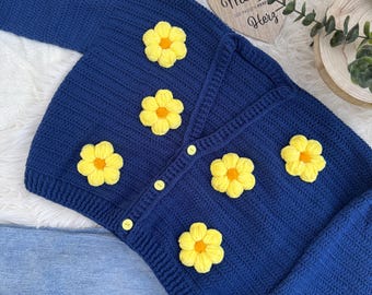 May include: A navy blue crochet cardigan adorned with yellow flower appliqués. The cardigan features a button-up front and long sleeves. The garment is displayed with a pair of blue jeans.