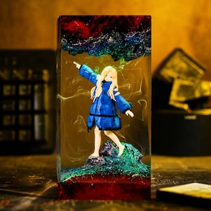 May include: A rectangular resin block containing a miniature figurine of a blonde person wearing a blue jacket. The figurine stands on a textured, multi-colored base, while swirling green and dark red resin patterns surround the figure.