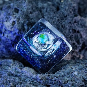 May include: A square-shaped resin keycap featuring a deep blue and black space-themed background with white speckles resembling stars. A small, spherical, blue and green planet sits at the center, surrounded by swirling white nebula-like patterns.