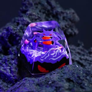 May include: A clear resin mechanical keyboard keycap featuring a purple Gengar character inside. The base of the keycap displays a dark purple and black landscape with swirling white patterns, while the character has red eyes and a red mouth.