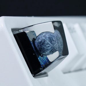 Könnte beinhalten: Eine individuelle mechanische Tastenkappe mit glänzendem, dunklem Harz-Finish und einem eingebetteten kugelförmigen Objekt, das ein detailliertes, strukturiertes blau-weißes Muster zeigt, das an eine Löwenzahnblüte erinnert, montiert auf einer weißen Tastatur.