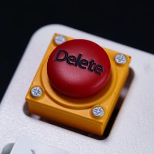 May include: A mechanical keyboard keycap featuring a circular red button with the word Delete printed in black. The button is mounted on a square yellow base secured by four silver-colored Phillips head screws, set into a white keyboard frame.