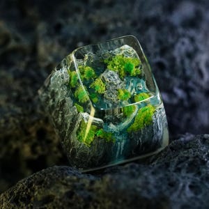 Puede incluir: Una tecla artesanal cuadrada que presenta un paisaje en miniatura encapsulado en resina transparente. El diseño incluye formaciones rocosas grises texturizadas, detalles de color verde vibrante similares al musgo y un elemento de agua azul translúcido.