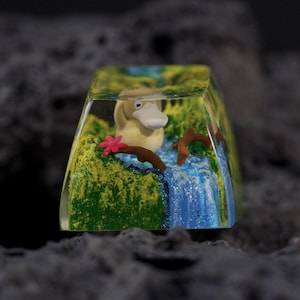 Puede incluir: Una tecla personalizada para teclado mecánico hecha de resina que presenta una figura en miniatura de una criatura amarilla similar a un pato, rodeada por un paisaje de musgo verde, ramas marrones, una pequeña flor rosa y una cascada azul.