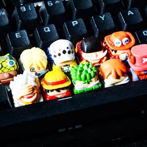以下が含まれることがあります： 黒いコンピューターキーボードの上に配置された、10個のカラフルなキャラクター型キーキャップのコレクション。各キーキャップには、異なる髪の色、帽子、表情を持つ詳細なミニチュアフィギュアが施されており、赤、緑、黄色のアクセントが特徴です。