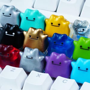 Könnte beinhalten: Eine Sammlung von elf individuellen mechanischen Tastaturkappen in Form von amorphen Blobs mit einfachen Gesichtern. Die Tastenkappen haben verschiedene Oberflächen, darunter metallisches Silber, mattes Schwarz, durchscheinendes Rot, leuchtendes Gelb, Blau, Lila und Grün.