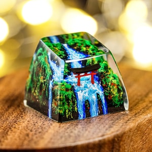 May include: A clear resin mechanical keyboard keycap featuring a detailed miniature landscape. The design depicts a vibrant blue waterfall cascading down between lush green cliffs, with a small red torii gate positioned in the centre of the flow.