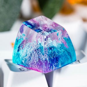 May include: A translucent resin mechanical keyboard keycap featuring a swirling internal design of vibrant purple, bright blue, and white pigments. The keycap has a smooth, glossy finish and sits atop a white keyboard key, showcasing a cloud-like pattern.
