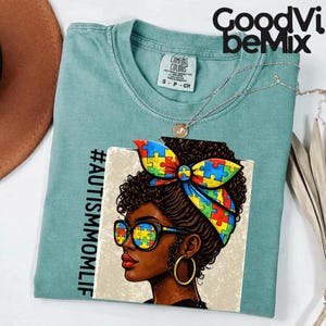 Puede incluir: Una camiseta Comfort Colors verde azulado con un gráfico de una mujer con una diadema y gafas con un patrón de piezas de rompecabezas. El texto "#AUTISMMOMLIF" está impreso verticalmente a la izquierda. Un collar plateado está drapeado sobre la camiseta.