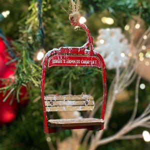 Könnte beinhalten: Roter Weihnachtsschmuck in Form eines Sessellifts. Der Schmuck trägt den Text "Santa's Express Chair Lift" und einen hölzernen Sitz. Er hängt an einem Weihnachtsbaum mit anderen Dekorationen und Lichtern.