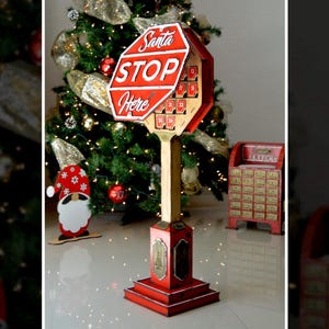 Könnte beinhalten: Rote und goldene Weihnachtsdekoration in Form eines Stoppschildes mit der Aufschrift "Santa Stop Here". Das Schild ist auf einem Holzpfosten und Sockel montiert. Ein Weihnachtsbaum und andere Dekorationen befinden sich im Hintergrund.