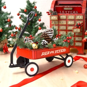 Op de afbeelding: Een feestelijke kerstscène met een rode wagen gevuld met dennenappels, groen en bessen. Op de wagen staat "Red Flyer". Kerstbomen met rode ornamenten en een rode brievenbus staan op de achtergrond.