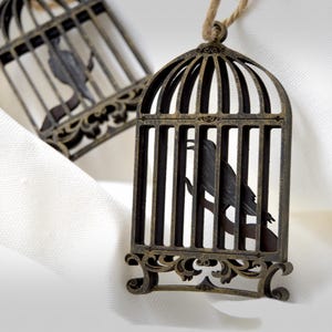 May include: Two decorative birdcage ornaments with a dark, antique bronze finish. Each cage features a silhouette of a bird perched on a branch. The ornaments hang from a natural twine loop, set against a white, textured background.