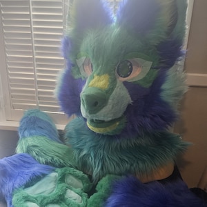 Op de afbeelding: Een dierenkopmasker van imitatiebont met blauwe, turquoise en limoengroene synthetische vacht, grote expressieve ogen en een bijpassende set poothandschoenen. Het masker heeft puntige oren en een snuit met een groene neus en turquoise zolen.