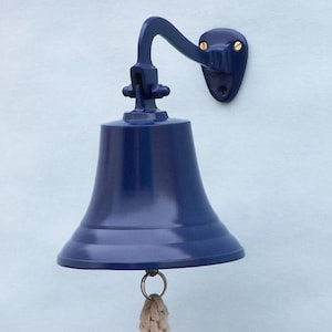May include: A navy blue nautical bell with a curved arm and mounting plate. The bell has a flared base and a rope pull. The bell is mounted on a light blue wall.