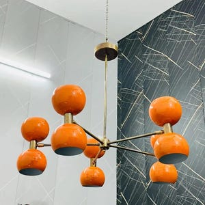 Könnte beinhalten: Ein Kronleuchter im Mid-Century-Modern-Stil mit acht orangefarbenen Kugelschirmen. Die Leuchte hat eine Messingoberfläche und ist von der Decke abgehängt. Die kugelförmigen Schirme sind glänzend und erzeugen eine Retro-Ästhetik.