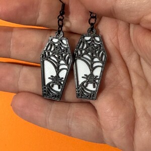 May include: Black coffin-shaped earrings with a white interior, a spider, and a spiderweb design. The earrings have black hooks and are held against an orange background.