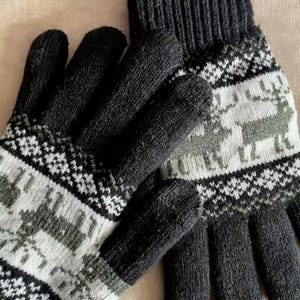 Może przedstawiać: Para czarnych dzianinowych rękawic z białym i zielonym geometrycznym wzorem i wzorem renifera. Rękawice wykonane są z grubego, ciepłego materiału, idealnego na zimną pogodę. Wzór jest widoczny na grzbiecie dłoni i na mankiecie.