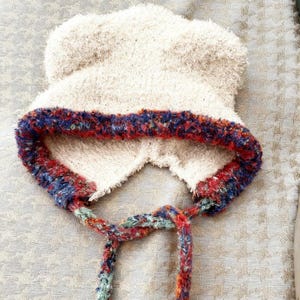 May include: A cream-coloured, fuzzy hat with a multi-coloured, textured trim in red, blue, and green. The hat has ear flaps and ties with a braided cord. The hat is made of a soft, plush material.