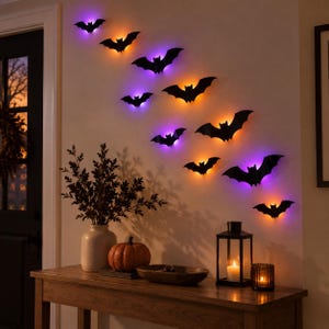 Peut inclure: Un ensemble de décorations murales en forme de chauves-souris noires disposées en diagonale, chacune rétroéclairée par des lumières LED violettes ou orange. Elles sont fixées sur un mur au-dessus d'une console en bois avec un vase.