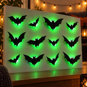 May include: A rectangular white board featuring twelve black bat-shaped cutouts arranged in a grid. Each bat is backlit with a vibrant green light, creating a glowing effect around the edges of the silhouettes against the white surface.