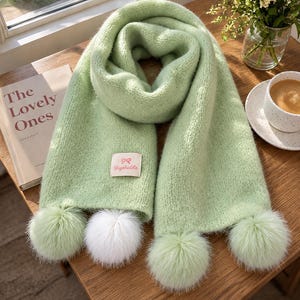 May include: A soft, light green knitted scarf featuring a rectangular fabric label with a pink bow icon and the text Uyetable. The scarf ends are decorated with three fluffy pom-poms, two in light green and one in white, creating a textured look.