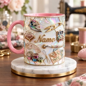 Może przedstawiać: Ceramiczny kubek z różowym uchwytem i wnętrzem, posiadający opalizującą białą powierzchnię zewnętrzną ozdobioną wypukłymi motywami krawieckimi, takimi jak żelazko, nożyczki, poduszeczka na szpilki, szpulki nici oraz napis Name złotą czcionką.