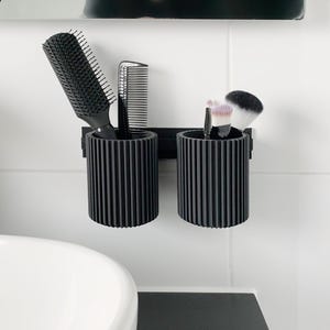 Könnte beinhalten: Zwei schwarze, geriffelte, zylindrische Halterungen an einer weißen gefliesten Wand. Einer enthält eine große schwarze Bürste und einen Kamm. Der andere enthält Make-up-Pinsel. Badezimmer-Organisation.