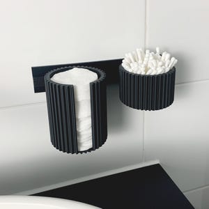 May include: A black, ribbed, wall-mounted organizer for bathroom essentials. The organizer features a cylindrical holder for cotton pads and a smaller, round container filled with cotton swabs. The design is modern and minimalist, suitable for a clean bathroom aesthetic.