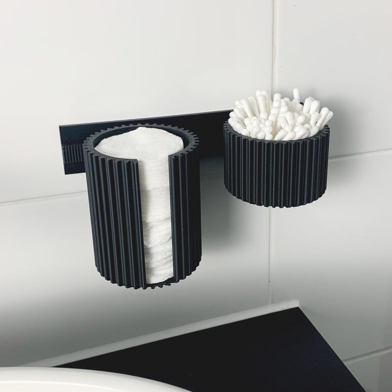 May include: A black, ribbed, wall-mounted organizer for bathroom essentials. The organizer features a cylindrical holder for cotton pads and a smaller, round container filled with cotton swabs. The design is modern and minimalist, suitable for a clean bathroom aesthetic.
