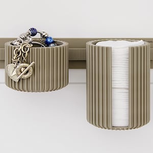 May include: A two-part, taupe-colored wall-mounted organizer. One section holds jewelry, the other holds cotton pads. The organizer has a ribbed design and is mounted on a white wall. The jewelry includes charms and beads.