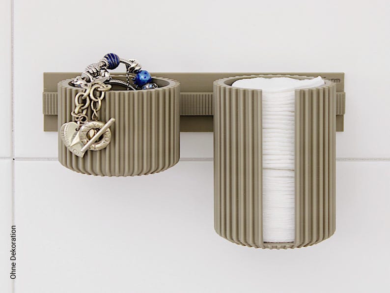 May include: A two-part, taupe-colored wall-mounted organizer. One section holds jewelry, the other holds cotton pads. The organizer has a ribbed design and is mounted on a white wall. The jewelry includes charms and beads.