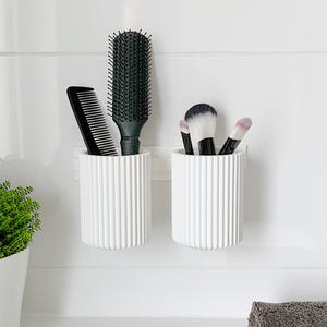 Könnte beinhalten: Zwei weiße, geriffelte, zylindrische Halterungen an einer weißen Wand. Einer enthält einen schwarzen Kamm und eine schwarze Haarbürste. Der andere enthält drei Make-up-Pinsel mit schwarzen Griffen und weißen und schwarzen Borsten. Links ist eine kleine grüne Pflanze zu sehen.