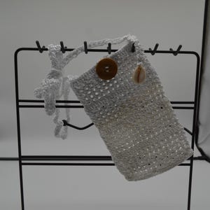 Puede incluir: Un bolso de ganchillo blanco con una correa larga, que presenta un único botón circular marrón en la parte superior y un pequeño dije de concha de color claro sujeto cerca de la abertura. El bolso tiene una textura de tejido abierto.