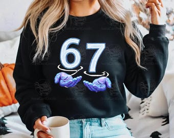 May include: Black sweatshirt with the number 67 in light blue, with a white outline. Below the numbers are two hands in purple, appearing to hold the numbers. The sweatshirt is a crew neck style.