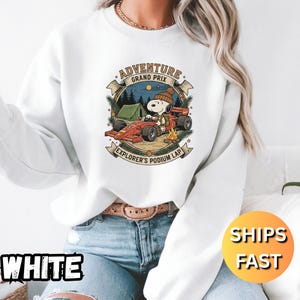 May include: White sweatshirt with a Snoopy graphic in a race car, text reads "Adventure Grand Prix Explorer's Podium Lap." The design includes a tent, campfire, and night sky. The word "WHITE" is printed at the bottom left.