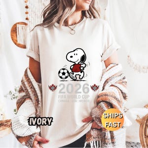 May include: Ivory-colored t-shirt featuring a cartoon Snoopy character kicking a soccer ball. The shirt has the text "2026 FIFA World Cup CANADA | USA | MEXICO" printed below the graphic. The shirt also has the word "IVORY" printed on the bottom left.