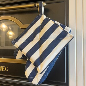 Puede incluir: Un par de manoplas a rayas azul marino y blancas colgadas de una barra de latón. Las manoplas son acolchadas y tienen un lazo para colgar. El fondo muestra un horno negro con la marca SMEG.