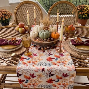 May include: A table setting featuring a white runner with a colourful autumn leaf print, a wooden centrepiece with assorted small pumpkins, dried wheat, and orange slices, flanked by tall taper candles in copper holders and amber-coloured glasses.