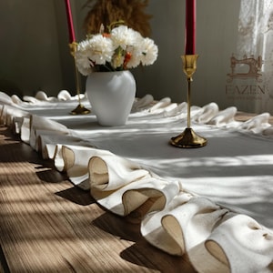 May include: A cream-coloured table runner with a ruffled edge is displayed on a wooden table. Two gold candle holders with red candles and a white vase with white and orange flowers are on the runner. The text "FAZEN PROFESSIONAL" is visible.