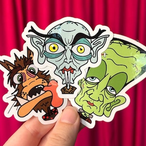 May include: A collection of three cartoon stickers featuring classic horror characters. The stickers include a werewolf, a vampire, and Frankenstein's monster, each with a unique, colorful design. The stickers are held against a pink background.