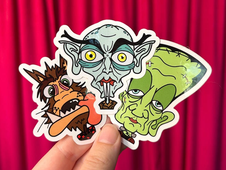 May include: A collection of three cartoon stickers featuring classic horror characters. The stickers include a werewolf, a vampire, and Frankenstein's monster, each with a unique, colorful design. The stickers are held against a pink background.