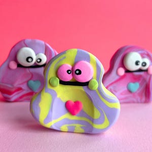 May include: Three colourful clay figures with large eyes and heart-shaped details. The central figure is yellow and purple, with two pink and purple figures on either side. Each has a heart and green accents.