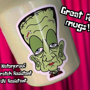 May include: A light green glittery mug with a sticker of a cartoon Frankenstein's monster. The sticker is green, white, black, and red. Text on the image says "Great for mugs!" and lists "Waterproof", "Scratch Resistant", and "UV Resistant".