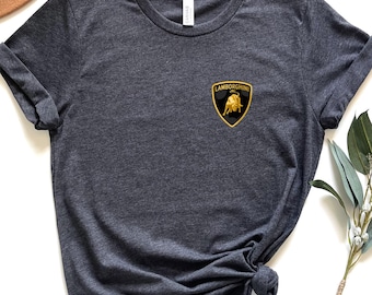 May include: Dark gray t-shirt with a small Lamborghini logo patch on the chest. The logo is a black shield with a gold bull and yellow border. The shirt is styled with rolled-up sleeves and a knot at the bottom.
